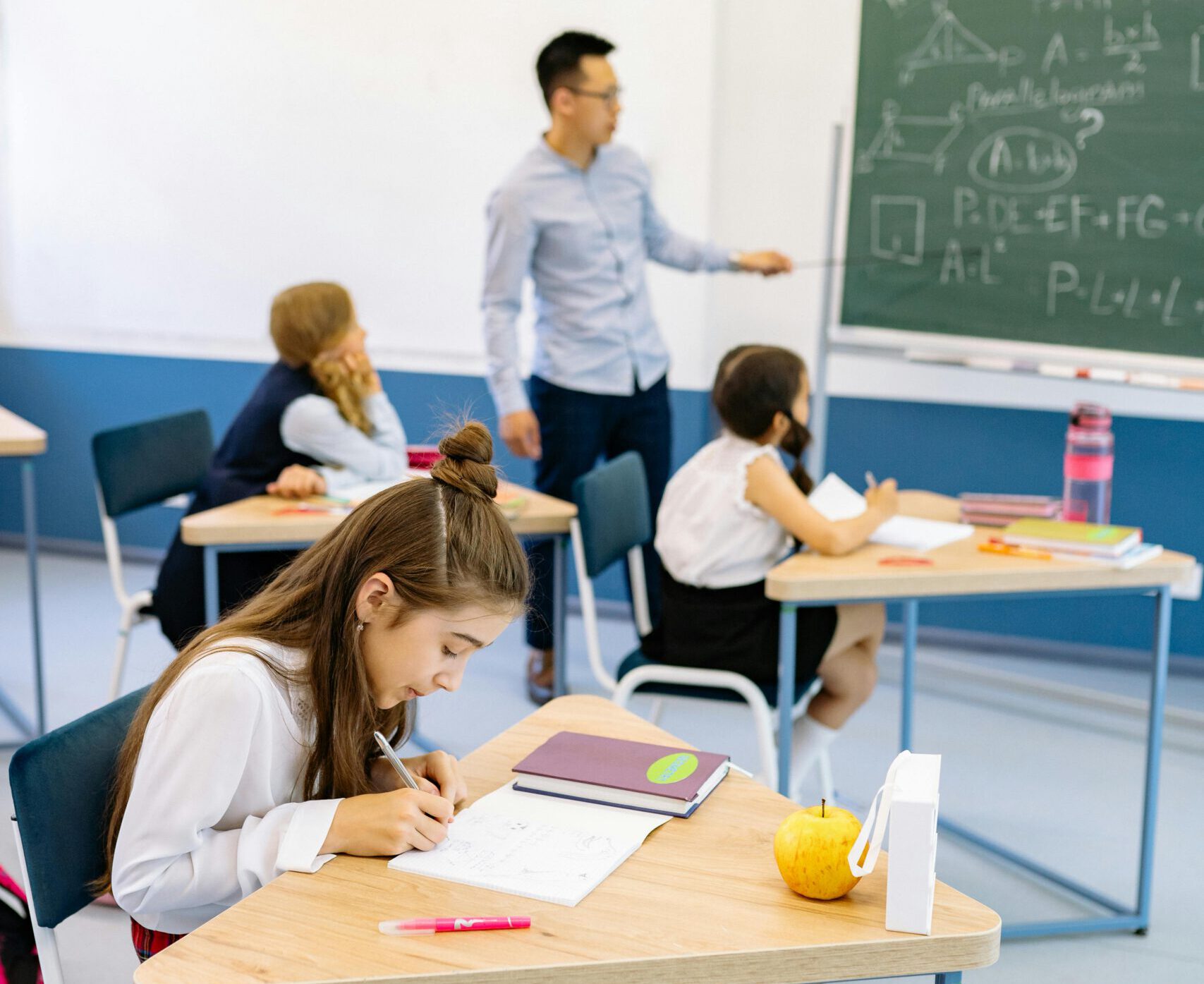 A teacher stands near a blackboard teaching students seated at desks in a classroom.
